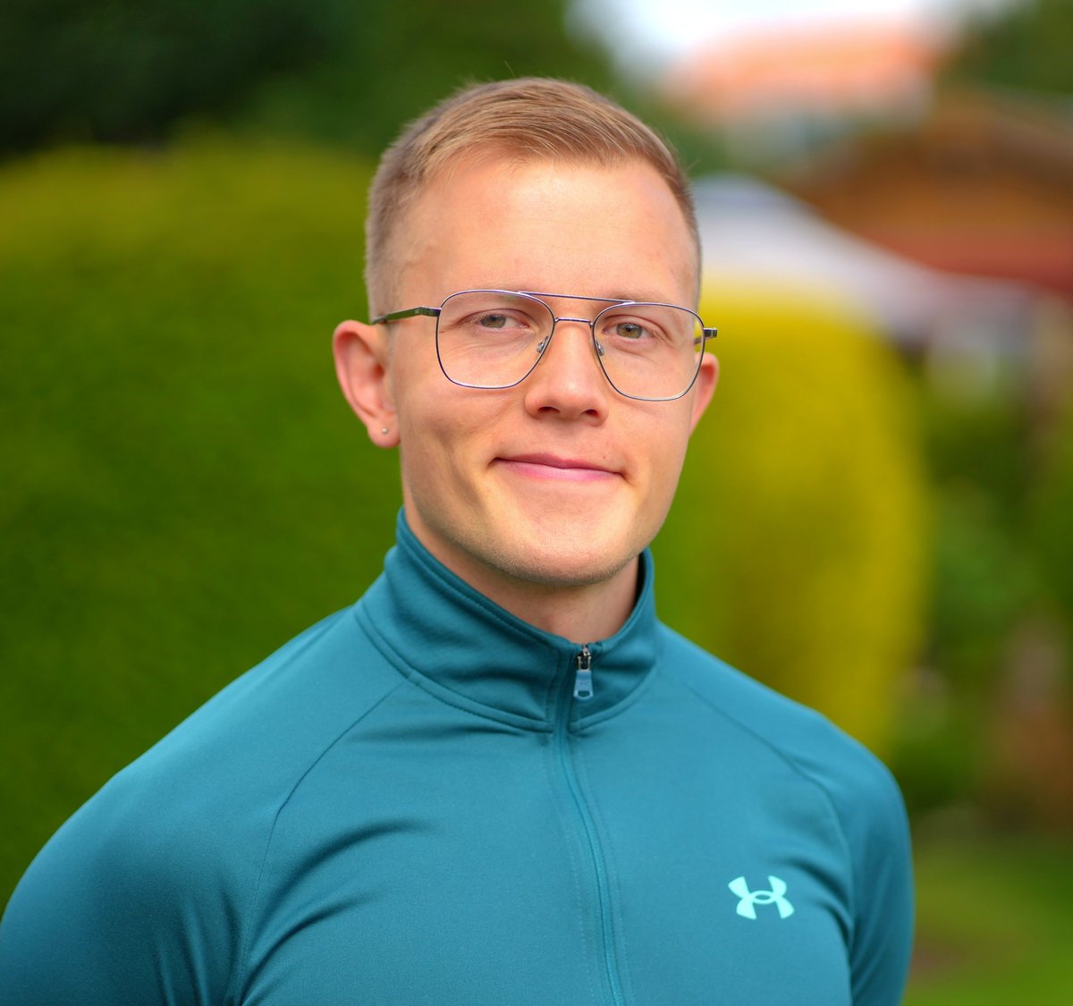 Young man wearing glasses and teal Under Armour jacket smiling outdoors with blurred green garden background